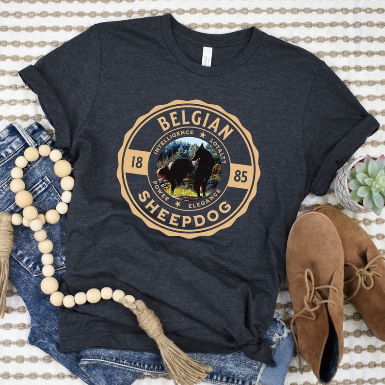 Op de afbeelding: Een donkergrijs T-shirt met een bruine en gouden cirkelvormige afbeelding met de tekst "Belgian Sheepdog" met een afbeelding van een zwarte en bruine hond in de cirkel. De afbeelding bevat ook de woorden "Intelligentie, Loyaliteit, Kracht, Elegantie" en de nummers "18" en "85".