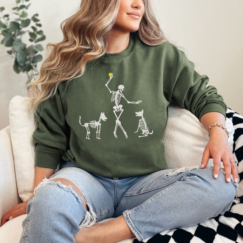 May include: A woman wearing a green sweatshirt with a white skeleton graphic of a person throwing a ball to two dogs. The skeleton is wearing a baseball cap and the dogs are sitting and waiting for the ball.