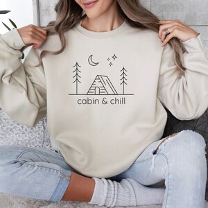 May include: A cream-colored sweatshirt with a black graphic of a cabin, trees, a moon, and stars. The text "cabin & chill" is printed below the graphic.