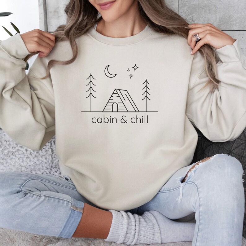 May include: A cream-colored sweatshirt with a black graphic of a cabin, trees, a moon, and stars. The text "cabin & chill" is printed below the graphic.