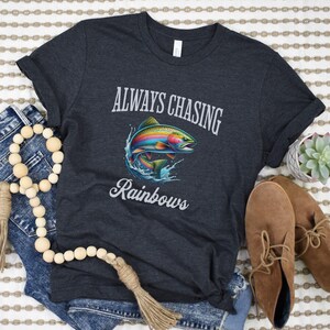 May include: A dark heather grey t-shirt with the text "ALWAYS CHASING RAINBOWS" printed in white. A rainbow trout fish is pictured below the text.