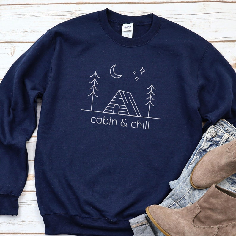 May include: A navy blue sweatshirt with a white outline of a cabin, trees, a moon, and stars. The text "cabin & chill" is printed below the image.