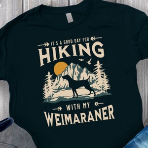 May include: A dark green t-shirt with a graphic design of a mountain range, a sunset, and a Weimaraner dog. The text on the shirt reads "It's a good day for hiking with my Weimaraner."