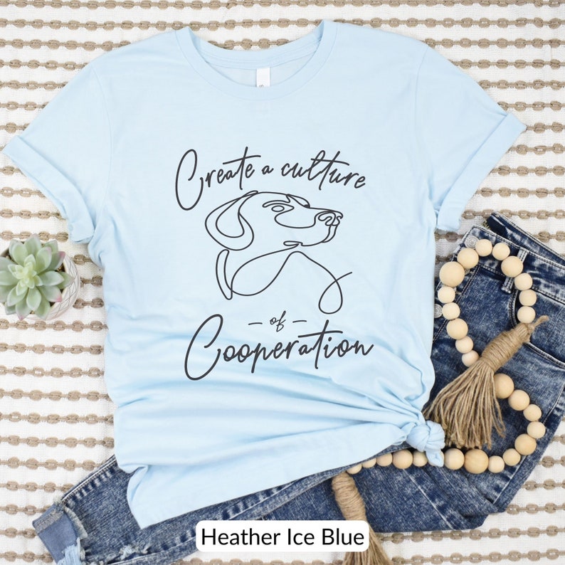 May include: Light blue t-shirt with a black line drawing of a dog's head and the text "Create a culture of Cooperation".