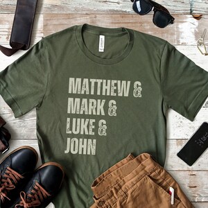 May include: Olive green t-shirt with the names Matthew, Mark, Luke, and John printed in a distressed, off-white font. The shirt is displayed with brown pants, black shoes, sunglasses, and a smartphone.