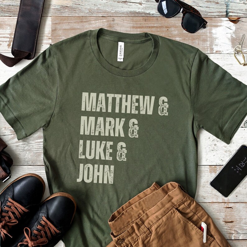 May include: Olive green t-shirt with the names Matthew, Mark, Luke, and John printed in a distressed, off-white font. The shirt is displayed with brown pants, black shoes, sunglasses, and a smartphone.