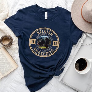 Op de afbeelding: Donkerblauw T-shirt met een beige cirkelvormige afbeelding met een Belgische herder en de tekst "BELGIAN SHEEPDOG" met de woorden "INTELLIGENCE", "LOYALTY", "POWER", "ELEGANCE" en de nummers "18" en "85" rond de cirkel.