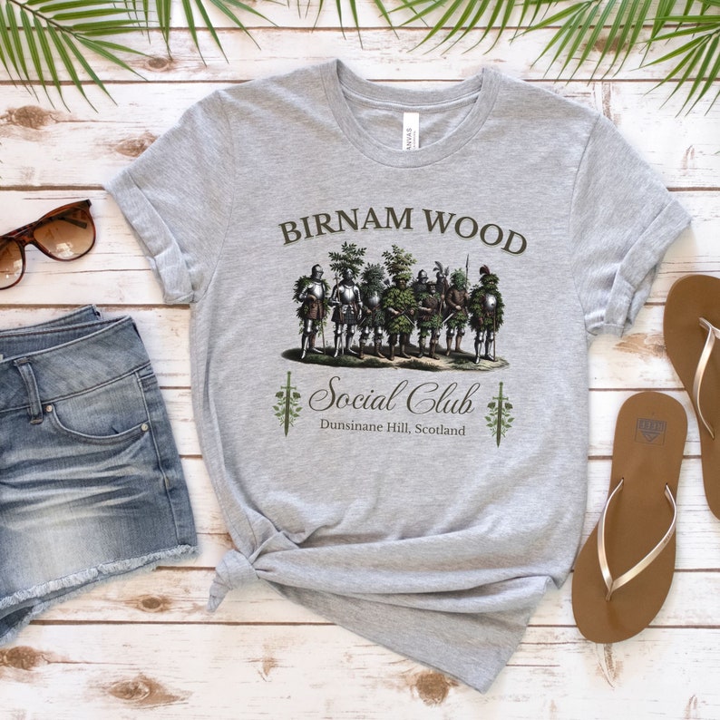 May include: A gray t-shirt with a graphic design of a group of men dressed as trees. The text "Birnam Wood Social Club" is printed on the shirt, along with the location "Dunsinane Hill, Scotland".