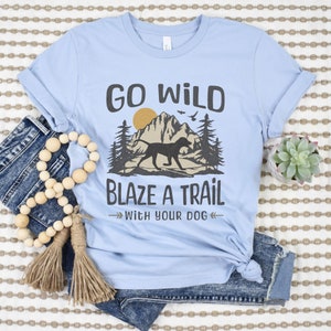 May include: Light blue t-shirt with a graphic design of a dog running through a mountain landscape with trees and the text "Go Wild Blaze a Trail with Your Dog".