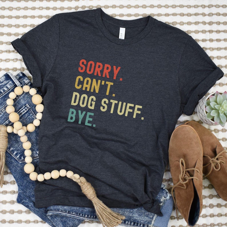 以下が含まれることがあります： 「SORRY. CAN'T. DOG STUFF. BYE.」とヴィンテージスタイルのグラフィックがプリントされたダークグレーのTシャツです
