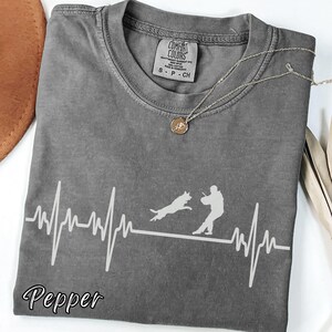 May include: A gray Comfort Colors t-shirt with a white graphic of a dog and person, and a heartbeat line design. The word "Pepper" is written in script at the bottom. A gold necklace with a pendant is on the shirt.