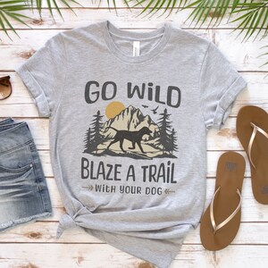 May include: Gray t-shirt with a graphic of a mountain range, a dog, and trees. The text "Go Wild Blaze a Trail with your Dog" is printed on the shirt.