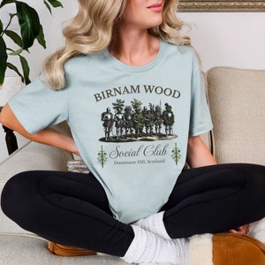 May include: A light blue t-shirt with a graphic of a group of people dressed as trees. The text "Birnam Wood Social Club" is printed on the shirt, along with the location "Dunsinane Hill, Scotland".