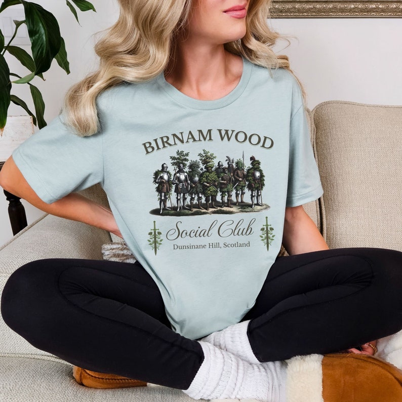 May include: A light blue t-shirt with a graphic of a group of people dressed as trees. The text "Birnam Wood Social Club" is printed on the shirt, along with the location "Dunsinane Hill, Scotland".