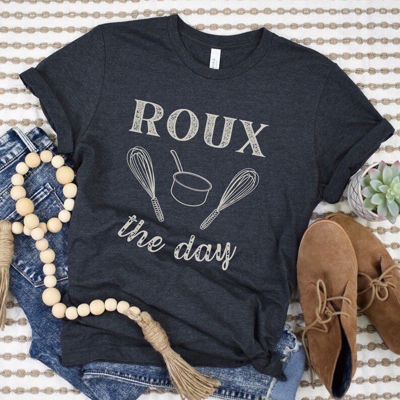 May include: A dark grey t-shirt with the text "ROUX the day" in white. The text is surrounded by two white whisk illustrations and a white pot illustration.