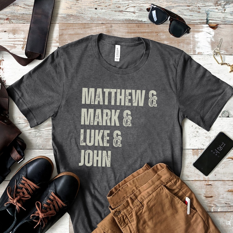 May include: Dark gray t-shirt with the names Matthew, Mark, Luke, and John printed in a distressed, off-white font. The shirt is displayed with brown pants, black shoes, sunglasses, and a smartphone.