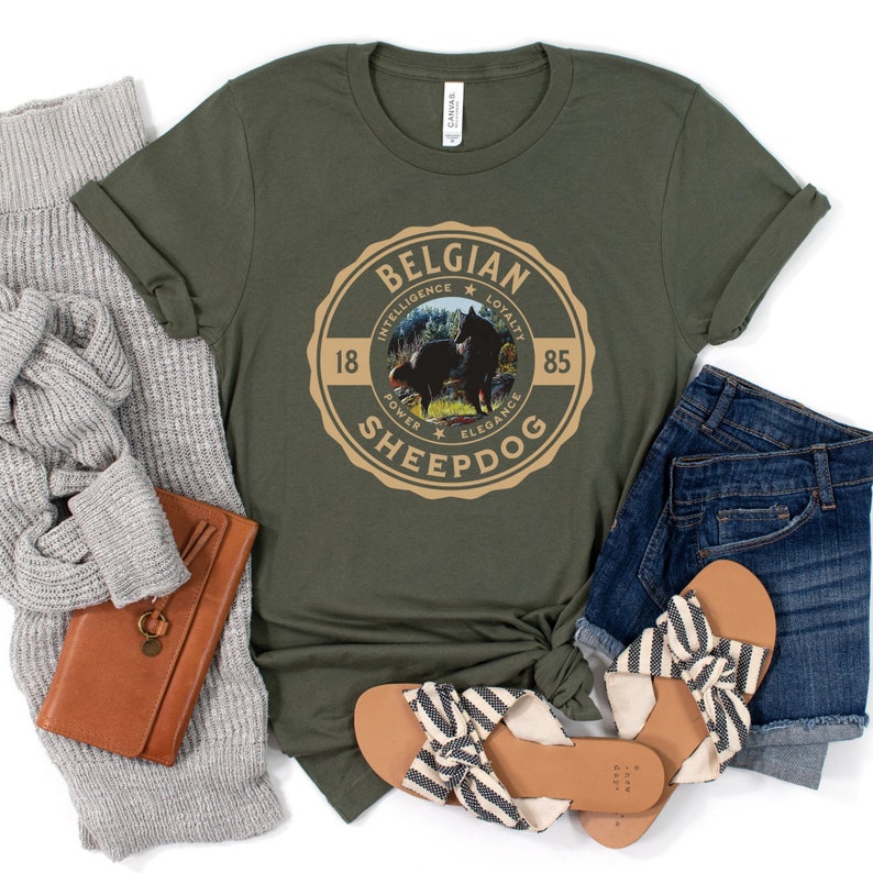Op de afbeelding: Een groen T-shirt met een bruine en beige ronde afbeelding waarop "Belgian Sheepdog" staat met een silhouet van twee zwarte honden in het midden. De afbeelding bevat ook de woorden "Intelligentie", "Loyaliteit", "Kracht", "Elegantie" en de nummers "18" en "85".