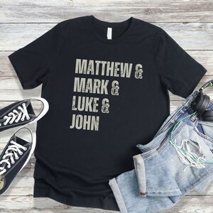 May include: Black t-shirt with the names "Matthew, Mark, Luke, and John" printed in a distressed, light gray font. The shirt is paired with black canvas sneakers, blue jeans, and headphones.