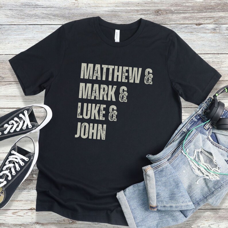 May include: Black t-shirt with the names "Matthew, Mark, Luke, and John" printed in a distressed, light gray font. The shirt is paired with black canvas sneakers, blue jeans, and headphones.