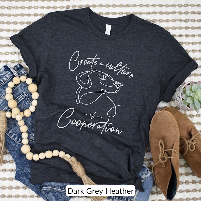 May include: Dark grey heather t-shirt with a white outline of a dog and the text "Create a culture of cooperation".