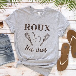 May include: A grey t-shirt with the text "ROUX the day" in brown lettering. The text is accompanied by a small pot and two whisks.