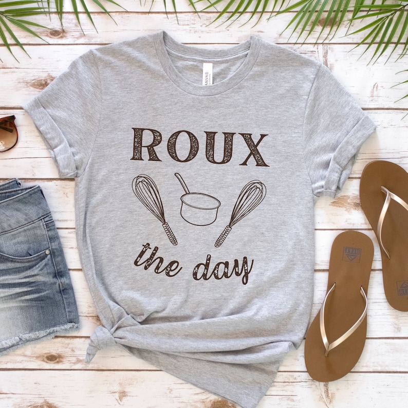 May include: A grey t-shirt with the text "ROUX the day" in brown lettering. The text is accompanied by a small pot and two whisks.