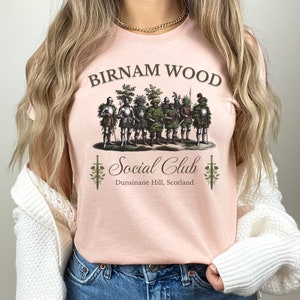 May include: A pink t-shirt with a graphic of a group of men dressed as trees. The text "Birnam Wood Social Club" is printed on the shirt, along with the location "Dunsinane Hill, Scotland".
