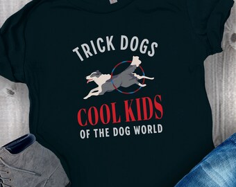 May include: A dark blue t-shirt with the text "Trick Dogs Cool Kids of the Dog World" in white and red lettering. A cartoon image of two dogs jumping through a hoop is in the center of the shirt.