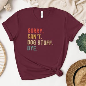 以下が含まれることがあります： レトロなフォントでカラフルなアクセントがついた「SORRY. CAN'T. DOG STUFF. BYE.」というテキストが書かれた、マルーンのTシャツです。
