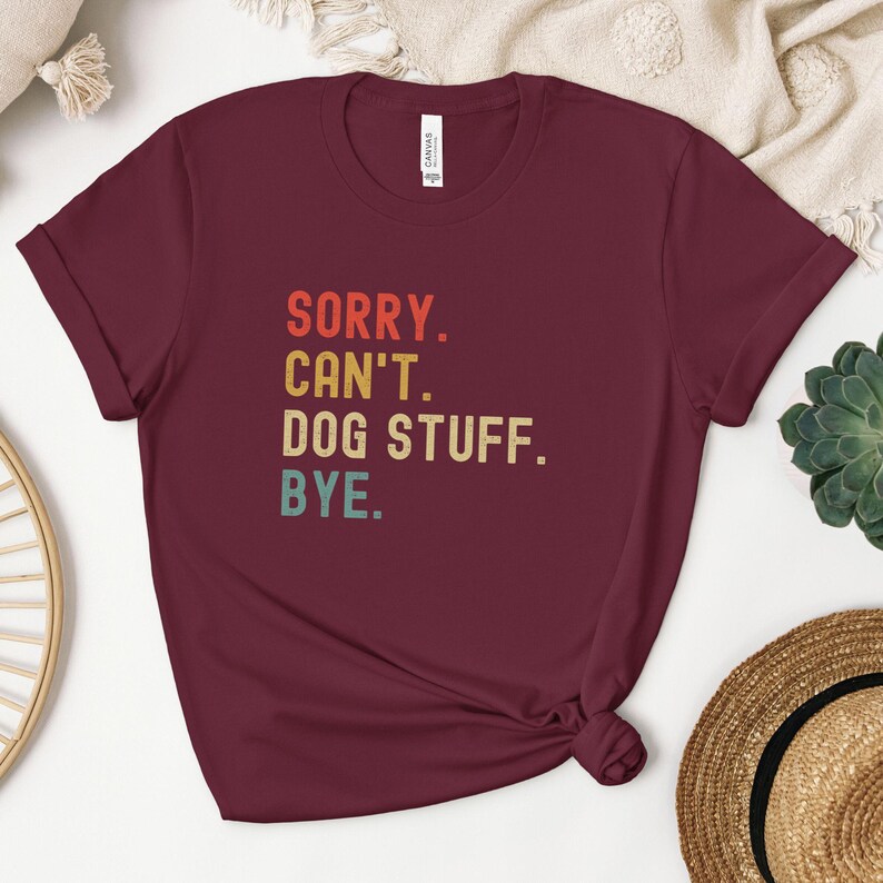 以下が含まれることがあります： レトロなフォントでカラフルなアクセントがついた「SORRY. CAN'T. DOG STUFF. BYE.」というテキストが書かれた、マルーンのTシャツです。