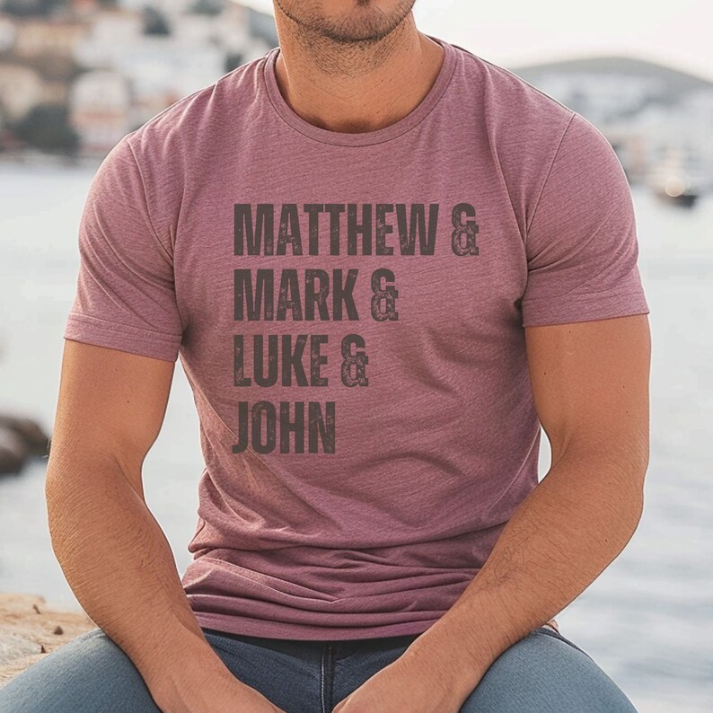 May include: A maroon crew neck t-shirt with the names "MATTHEW & MARK & LUKE & JOHN" printed in dark gray. The shirt is made of a soft material and is suitable for casual wear.