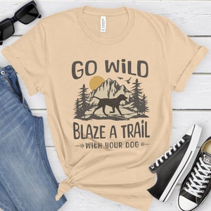 May include: Beige t-shirt with a graphic design of a mountain range, a dog, and the text "Go Wild Blaze a Trail With Your Dog".