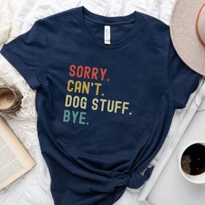 以下が含まれることがあります： レトロなフォントでカラフルなアクセントがついた「SORRY. CAN'T. DOG STUFF. BYE.」というテキストが書かれたネイビーブルーのTシャツです。
