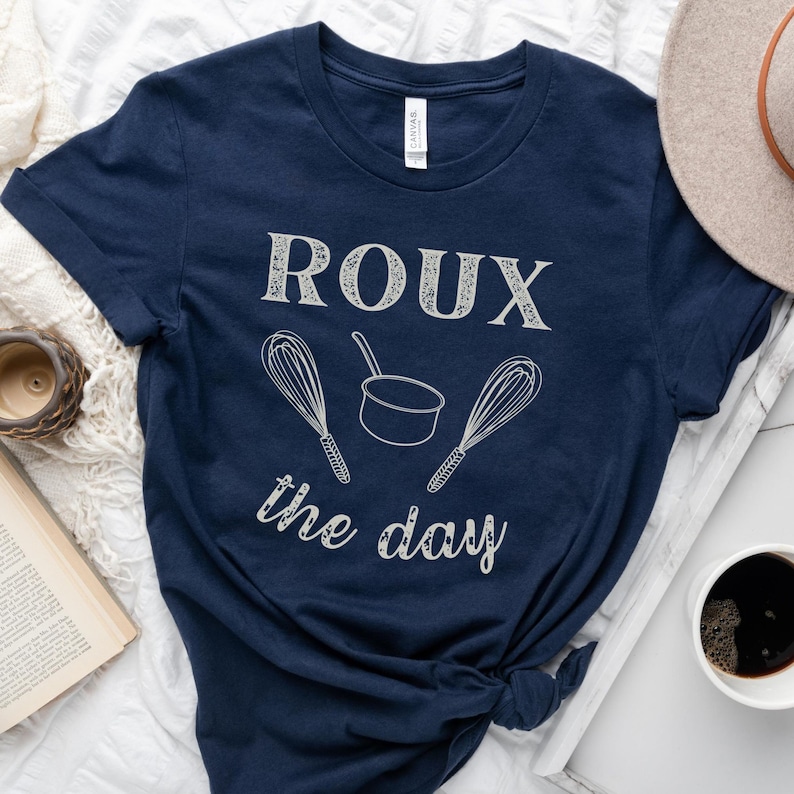 May include: A navy blue t-shirt with the text "ROUX the day" in white. The text is surrounded by two white whisk illustrations and a white pot illustration.