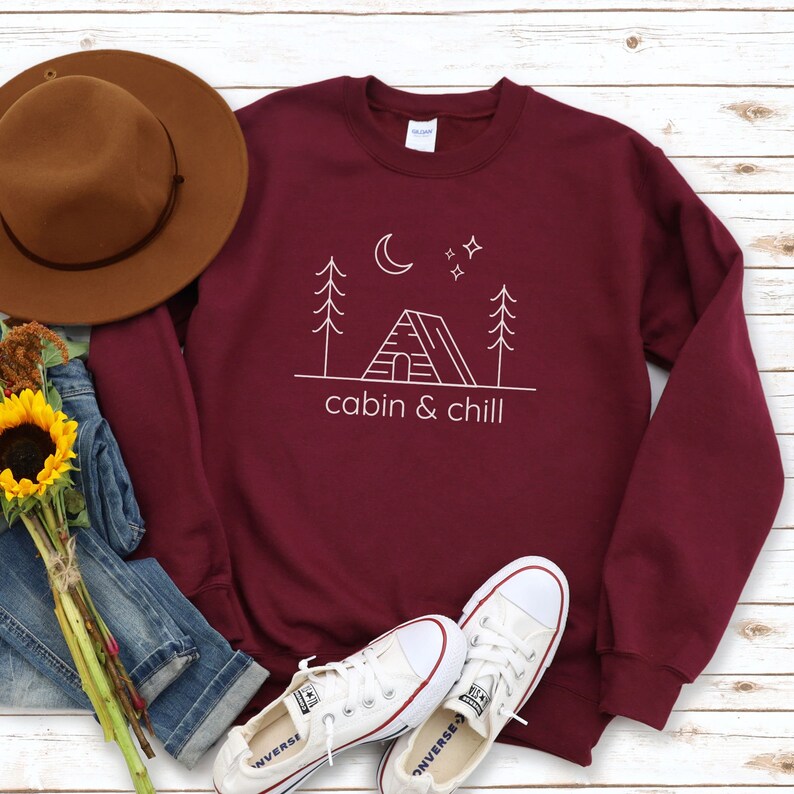 May include: A maroon sweatshirt with a white graphic design of a cabin, trees, a crescent moon, and stars. The text "cabin & chill" is printed below the graphic.