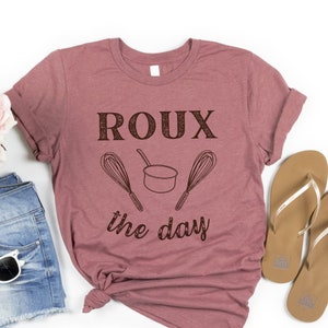 May include: A mauve t-shirt with the text "ROUX the day" in brown lettering. The text is accompanied by a small pot and two whisks.
