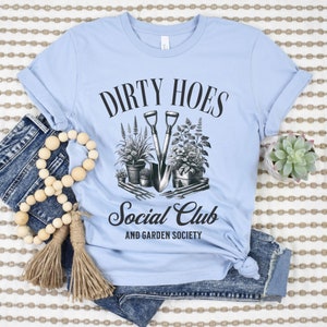 May include: Light blue t-shirt with black text that reads "Dirty Hoes Social Club and Garden Society". The text is surrounded by a black circle and an illustration of gardening tools and plants.