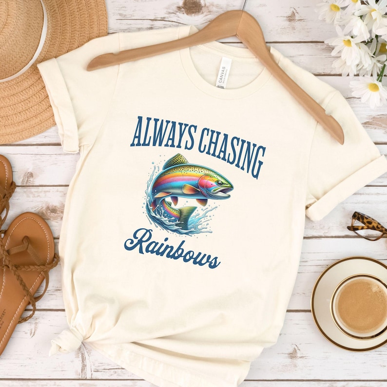 May include: A white t-shirt with the text "ALWAYS CHASING RAINBOWS" in blue lettering. A rainbow trout jumps out of the water, surrounded by blue waves.