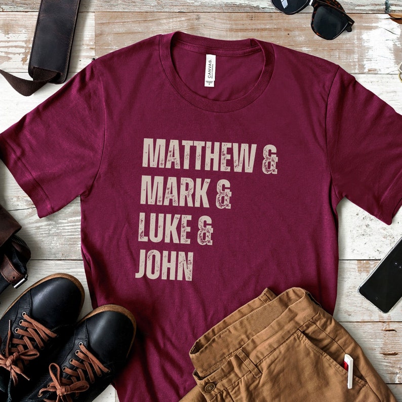 May include: A maroon t-shirt with the names Matthew, Mark, Luke, and John printed in a distressed, off-white font. The shirt is displayed with black leather shoes, brown pants, and sunglasses on a wooden surface.
