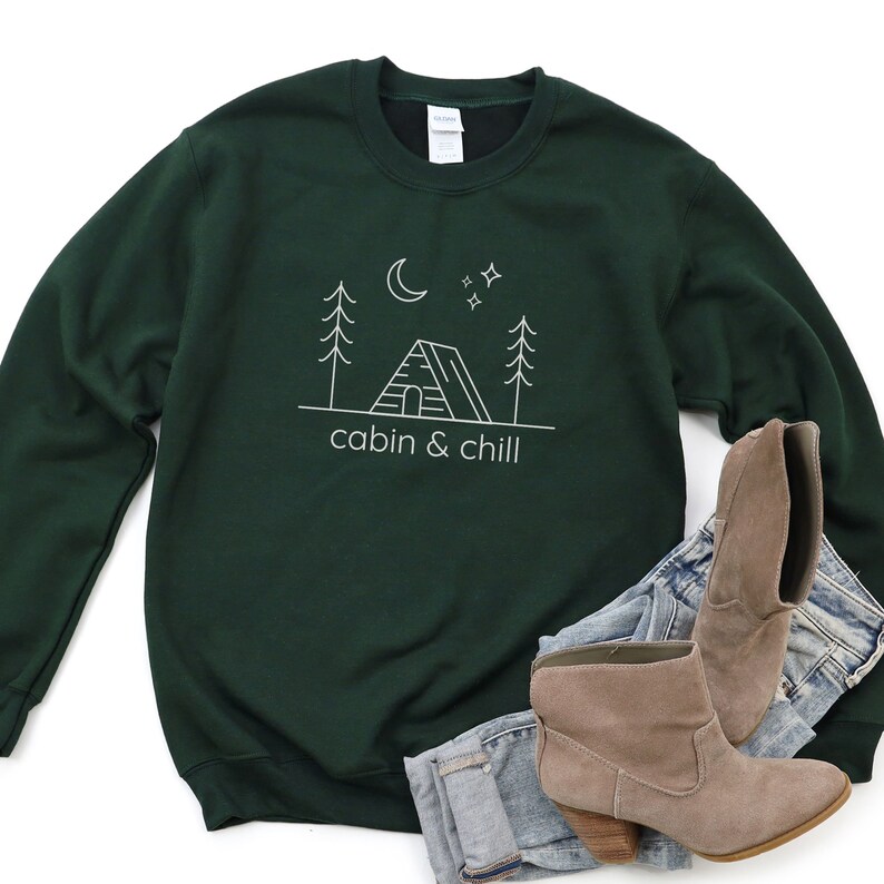 May include: A dark green sweatshirt with a white outline of a cabin, trees, a moon, and stars. The text "cabin & chill" is printed below the image.