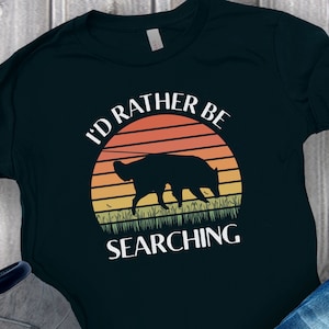 May include: Black t-shirt with a graphic design featuring a silhouette of a dog in front of a sunset. The text reads "I'D RATHER BE SEARCHING" in white, arched above and below the dog illustration. The design includes orange, yellow, and green colors.