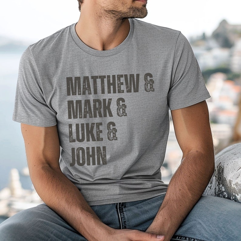 May include: A gray t-shirt with the names "MATTHEW", "MARK", "LUKE", and "JOHN" printed in a distressed, dark gray font. The shirt is a crew neck style.