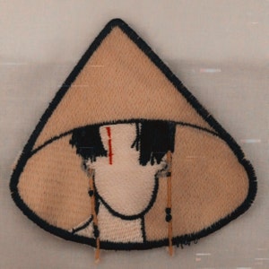 May include: A brown embroidered patch of a person wearing a conical hat with a red line on their forehead. The person has black hair and is wearing a white shirt. The patch is surrounded by a black outline.