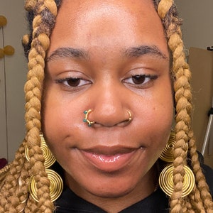 May include: A person with long, braided hair and gold jewellery. The person wears a nose ring with turquoise accents and large, circular earrings. The skin tone is warm, and the overall aesthetic is stylish.