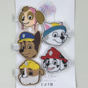 May include: A set of six embroidered hair clips featuring cartoon dog faces. The clips include characters in various colors, such as brown, blue, yellow, and red, attached to white ribbon. The clips are attached to a white card.