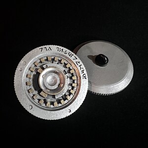 May include: Two-piece, silver-colored metal object with a circular design. One piece features a complex inner mechanism with gold-colored accents and etched text. The other piece is a simple disc with a black knob.