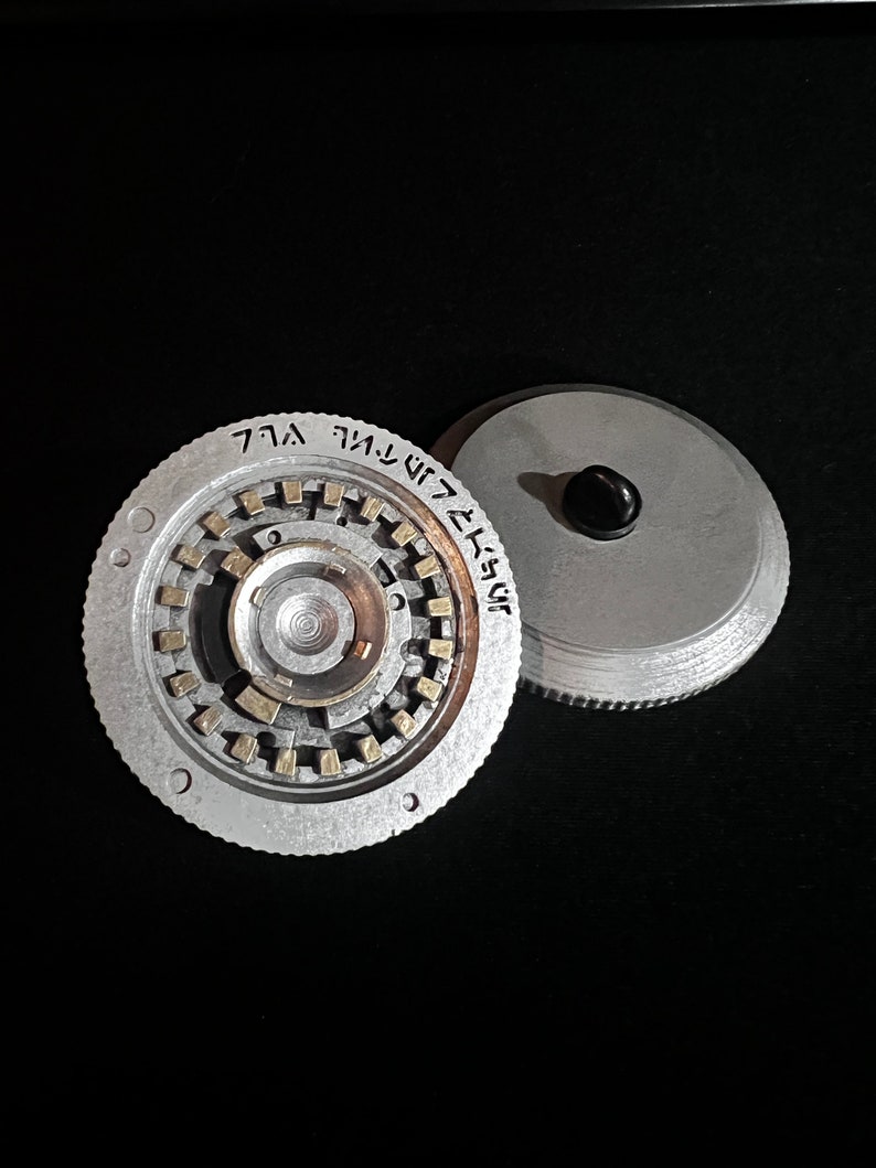 May include: Two-piece, silver-colored metal object with a circular design. One piece features a complex inner mechanism with gold-colored accents and etched text. The other piece is a simple disc with a black knob.