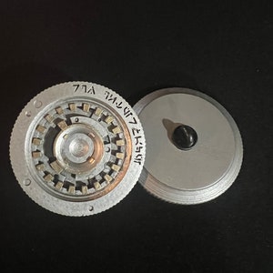 May include: Two-piece, silver-colored metal object with a circular design. One piece has a central mechanism with small, rectangular elements and etched text. The other piece is a plain, circular lid with a black knob. The objects are on a black surface.