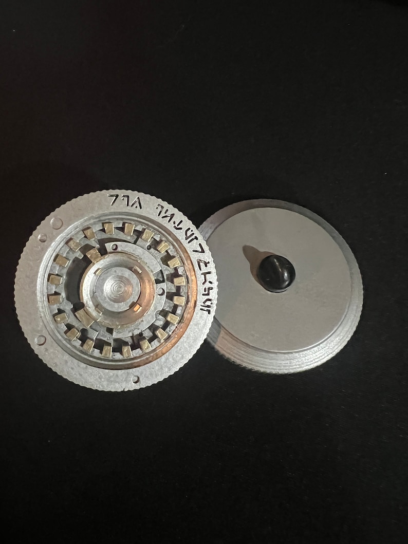 May include: Two-piece, silver-colored metal object with a circular design. One piece has a central mechanism with small, rectangular elements and etched text. The other piece is a plain, circular lid with a black knob. The objects are on a black surface.
