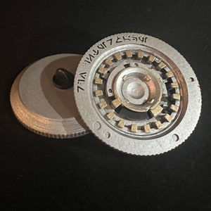May include: A silver-colored, circular metal object with a complex inner mechanism and gold-colored accents. The object is partially disassembled, revealing the intricate internal design. The rim features etched text. The background is black.